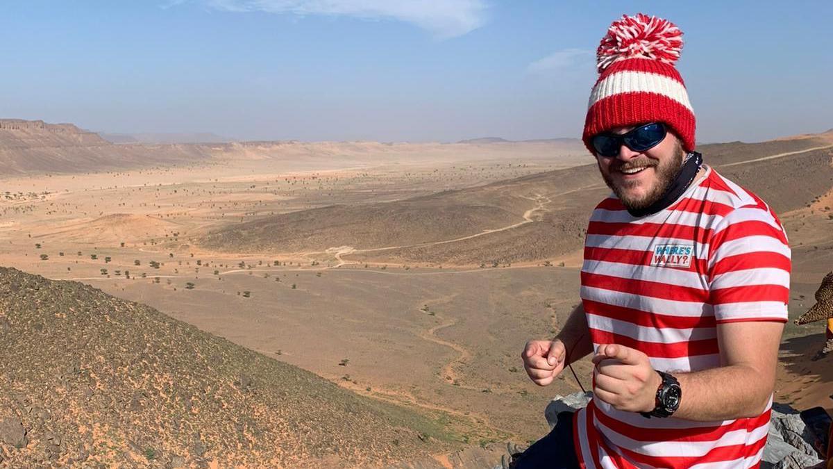 A man smiling and pointing at the camera with a desert in the background. He is wearing black sunglasses, a red and white striped T-shirt with the words "Where's Wally?" on and a red and white striped bobble hat.