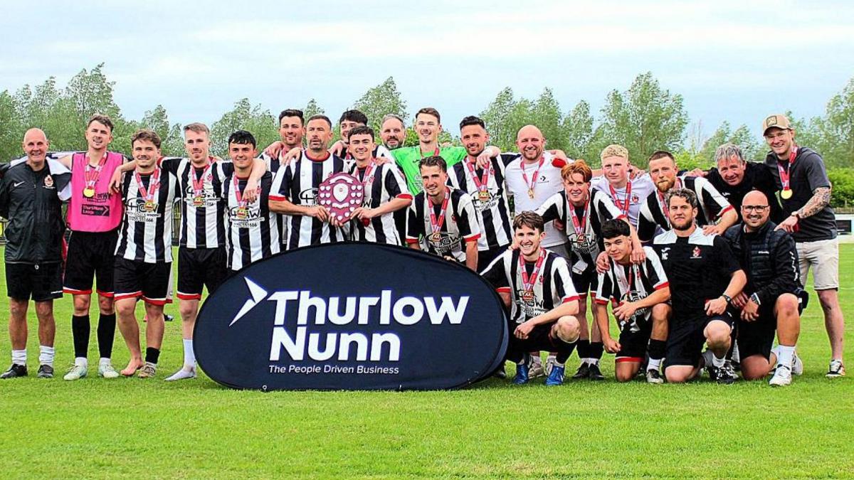 Harwich & Parkeston FC are being crowned champions of the league. They are crowding together in black and white striped shirts. They are on the green pitch with trees in the background. Two players at the front hold a shield.