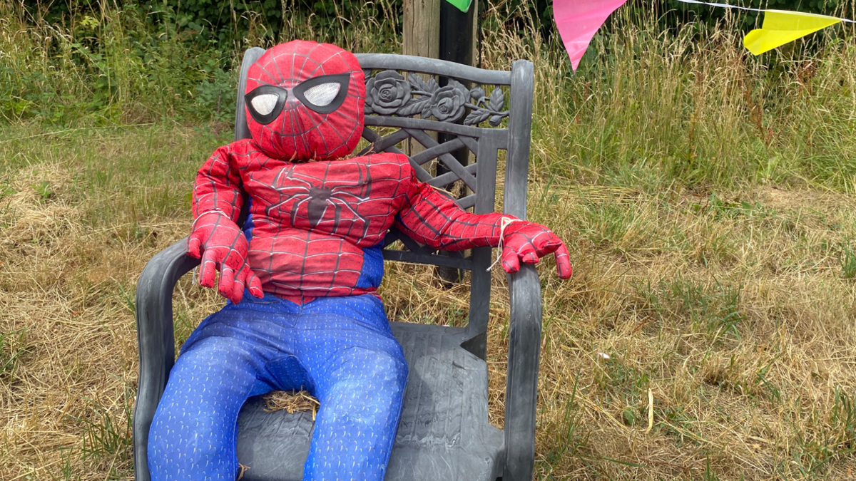 A scarecrow sitting on a wooden chair looks like spiderman. It has blue pants and the iconic black and red mask and top. 