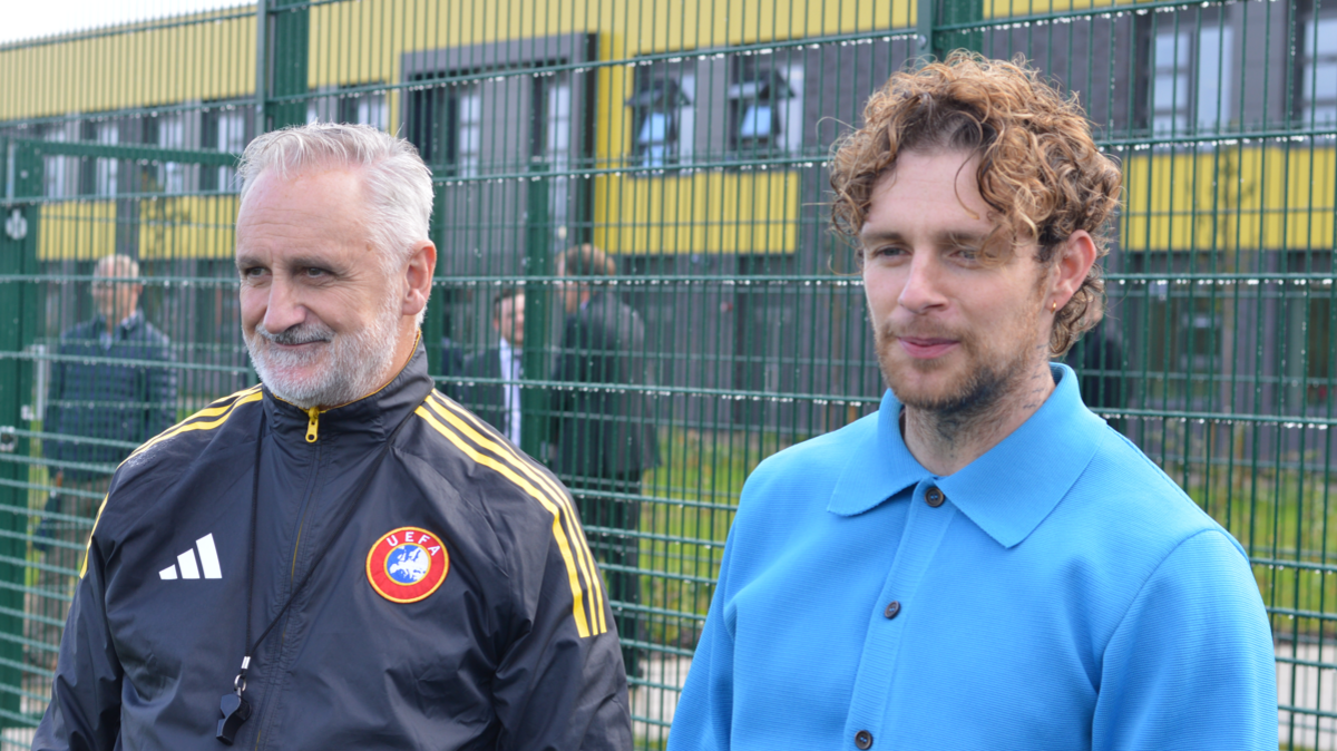 Tom Grennan pictured at the school new 3G sports pitch. He wears a bright blue long sleeve shirt with black buttons. He has curly hair, a gold ear piercing and a tattoo on his neck. He is stood to the right of a grey haired man wearing a navy blue tracksuit top with a round UEFA badge on. 