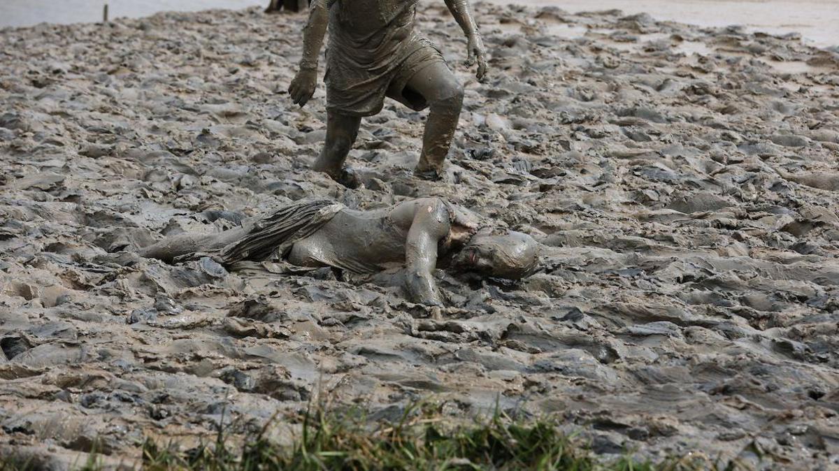 A woman lies face down in the mud, seemingly struggling to move forward. Another figure is knee-deep in mud behind her.