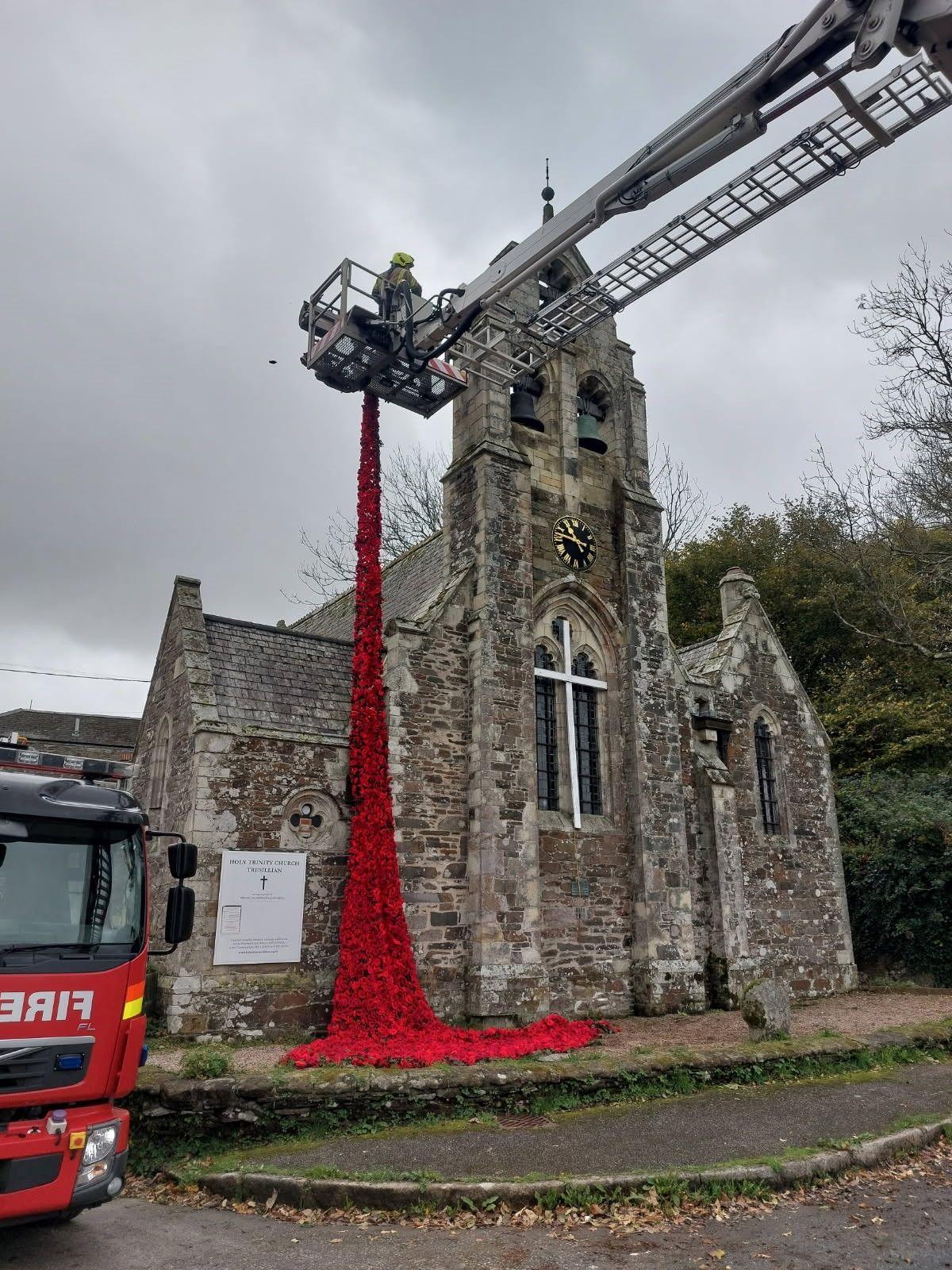 A firefighter in a cherry picker is seen draping the red poppy display on to a stone church building. Part of a fire engine is to the left of the image.