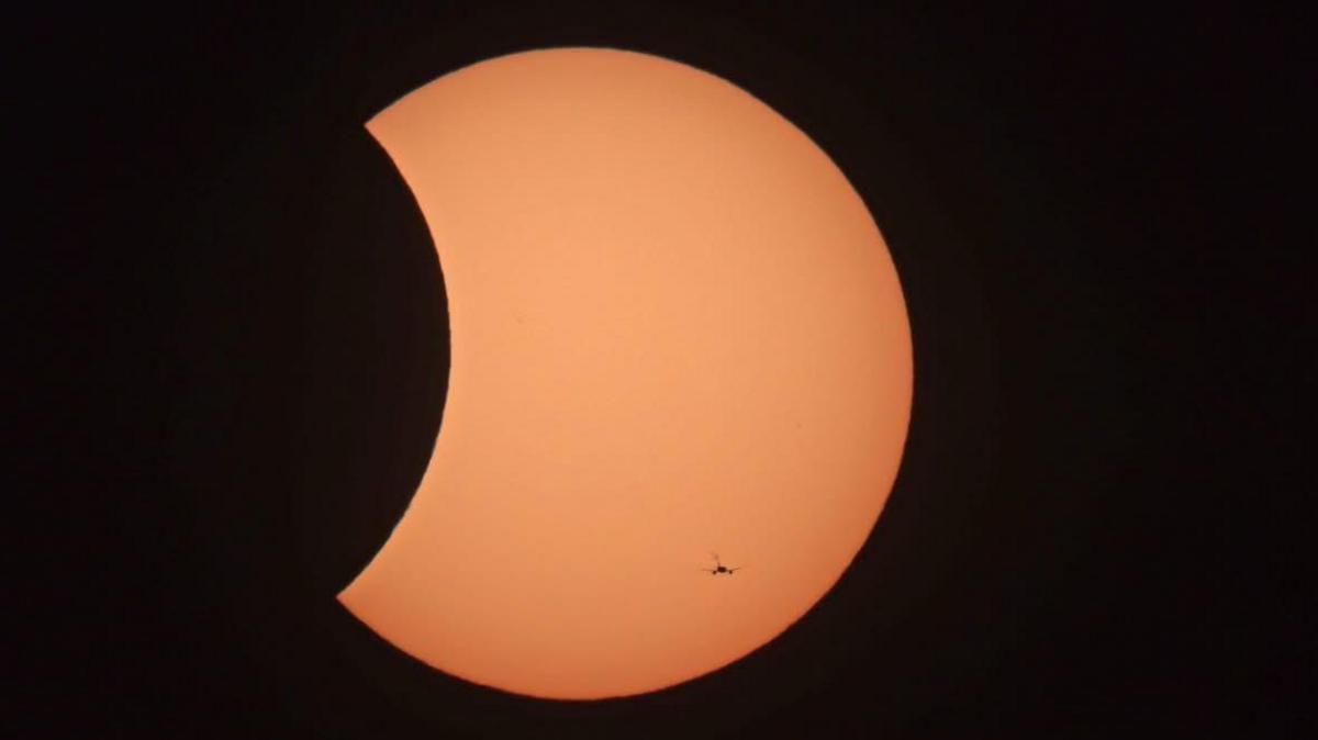A close-up photograph shows the Sun partially covered by the Moon during the eclipse in Sunbury-on-Thames, Surrey. The Sun appears as a large orange disc with a curved section missing, while a small helicopter silhouette can be seen flying across the face of the Sun.