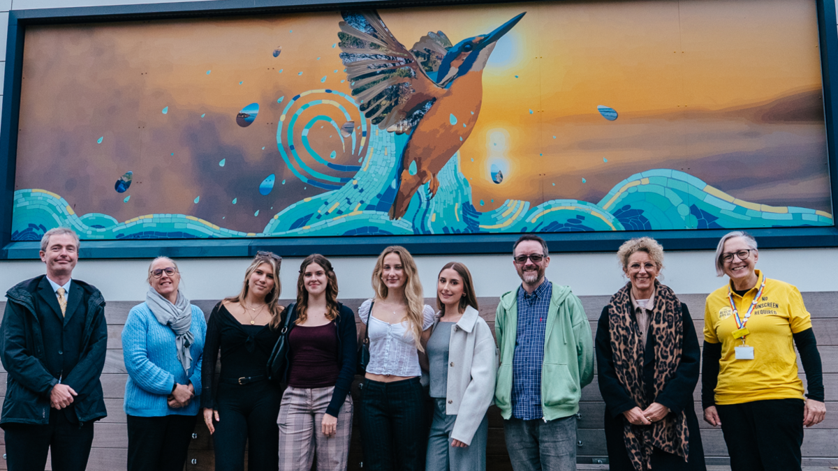A long line of nine men and women posing in front of a mural depicting a flying kingfisher set against an orange sunset. The bird is emerging from water and the droplets contain photos of locations, although the detail is difficult to make out.