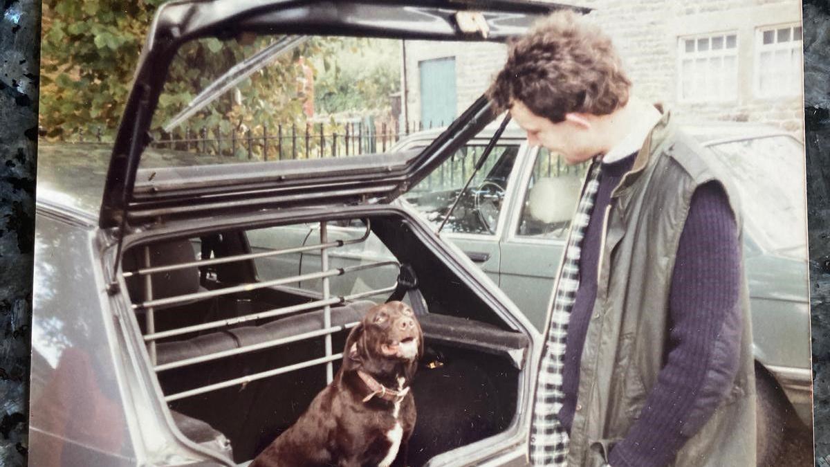 An old photo that is slightly lacking in vibrancy of Christopher. He is standing at the back of an old car which has its boot open. Sitting in side the booth is a brown dog. Christopher is looking and smiling at the dog. He wears a green sleeveless jacket with a blue jumper underneath. The dog has a brown collar around its neck.