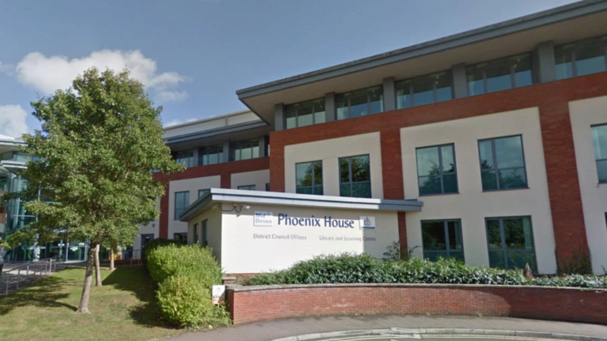 Phoenix House district council offices in Tiverton. A council building made of white and red brick. It has sets of four windows positions in a square and a small outbuilding with the logo written on it. There is a tree to the left of the image. 