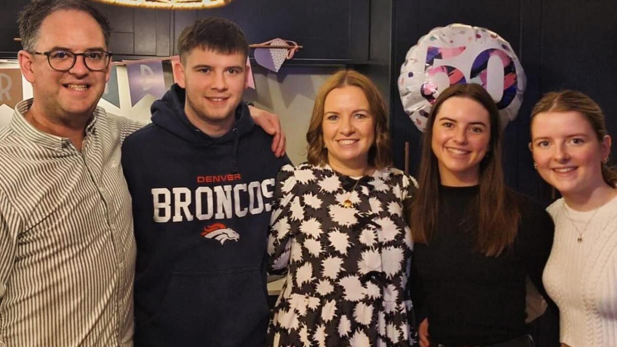 Lucy pictured linking arms with her family. 
Nicola King is wearing a white patterned shirt and Lucy is wearing a black top. Thomas King, Lucy's brother, is wearing a Denver Bronco's hoodie and their father, Richard is in a striped shirt. Lucy's sister, Olivia, is wearing a white top and necklace.  There is a balloon with the number 50 floating behind them.