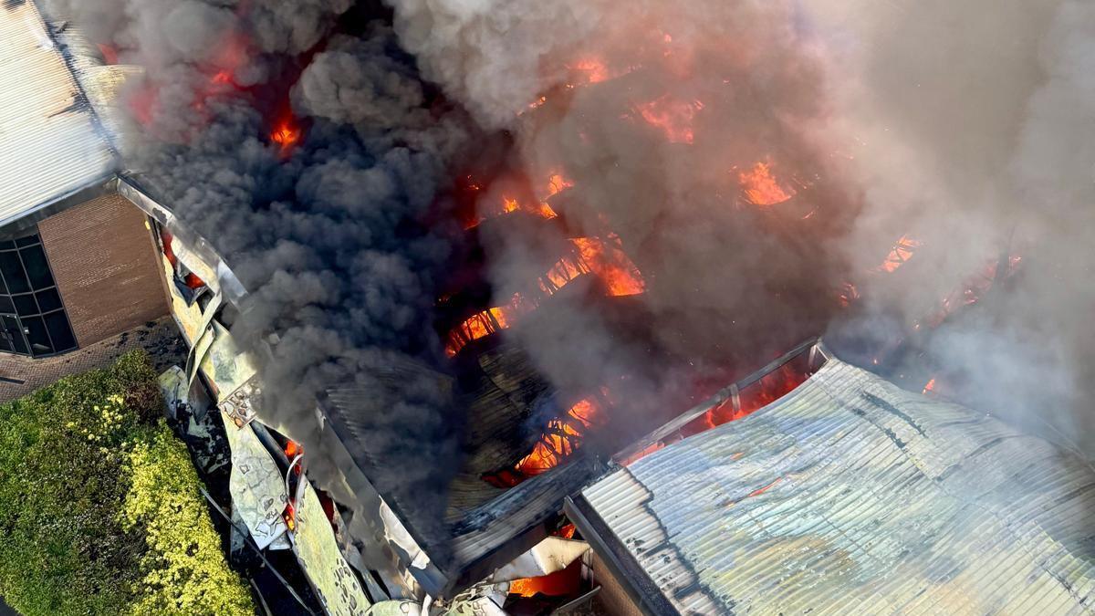 An aerial picture taken above the burning warehouse. Parts of the roof have collapsed and the parts that remain are charred black. Large flames can be seen through the gaps in the roof and black smoke is pouring out.