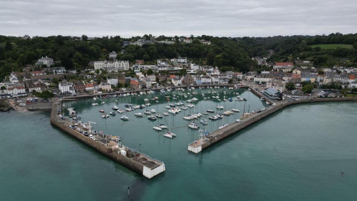 An ariel view of St Aubin in Jersey at high tide. There are boats in harbour and there are house surrounding the harbour. In between the buildings is green trees and green hills in the distance. 