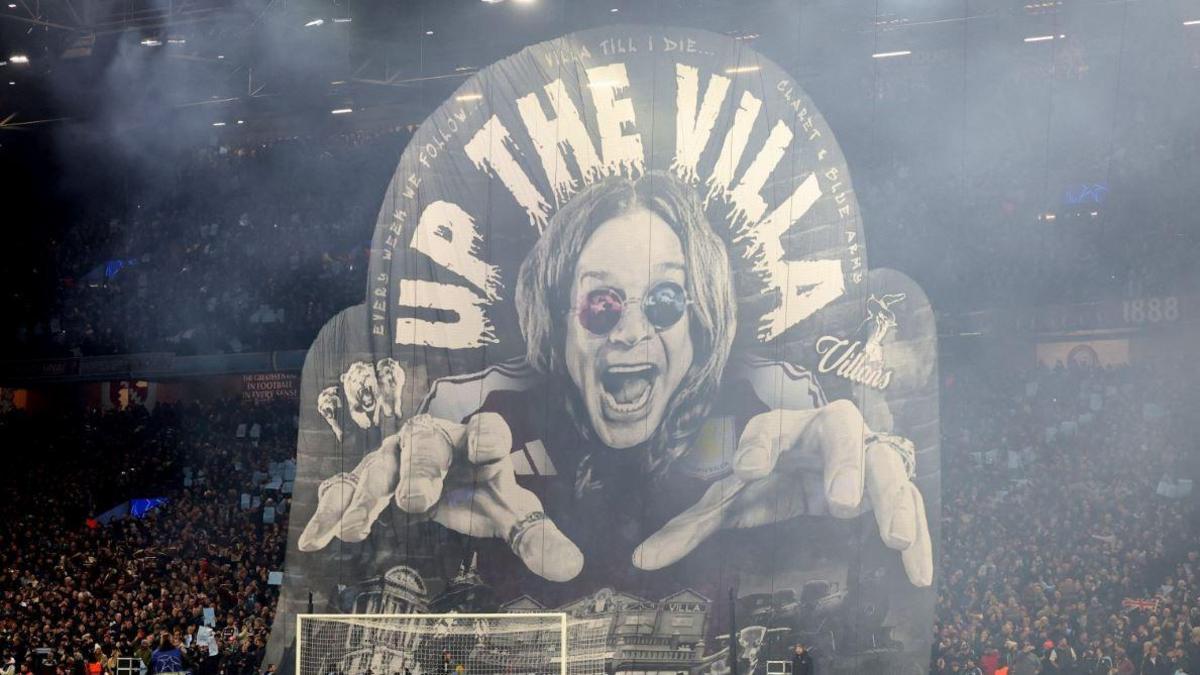 A giant flag black and white flag behind a goal post at Aston Villa featuring Ozzy Osbourne with an excited expression reaching out with his hands, with "Up the Villa" printed on it. The flag extends metres into the air, with football fans in the stands behind.