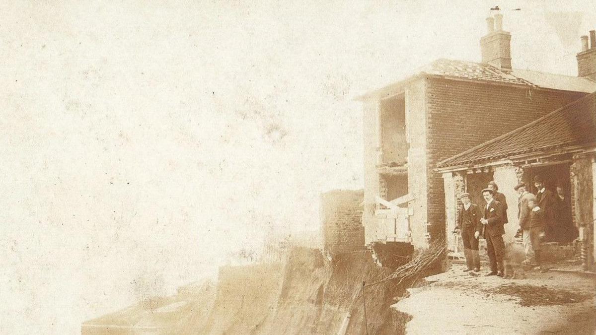 An aged image of a group of men standing and sitting in front of a building on an eroded coastline. 