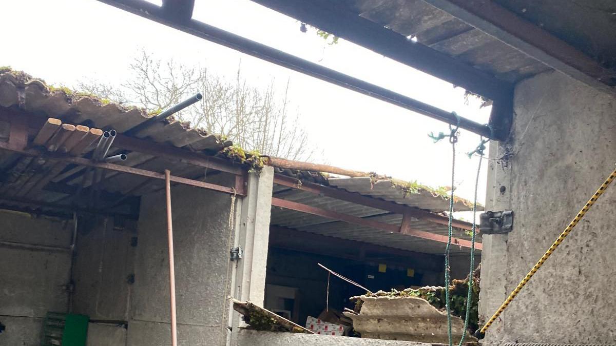 A photo from inside a garage showing a large gap in the corrugated roof and a hole in the dividing wall.