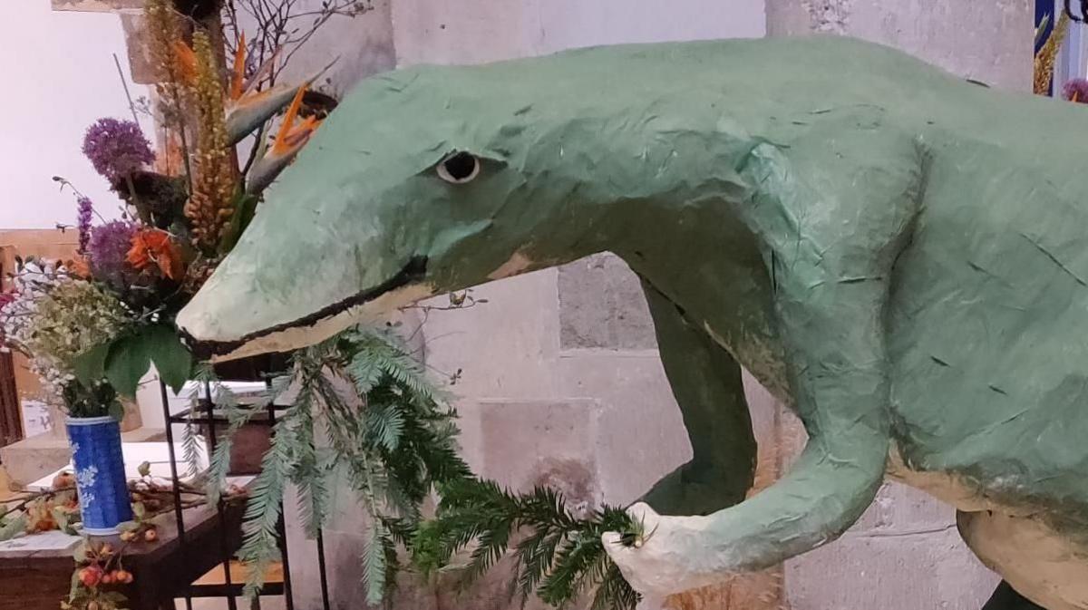 A large green papier mache dinosaur appears to be chewing a fern