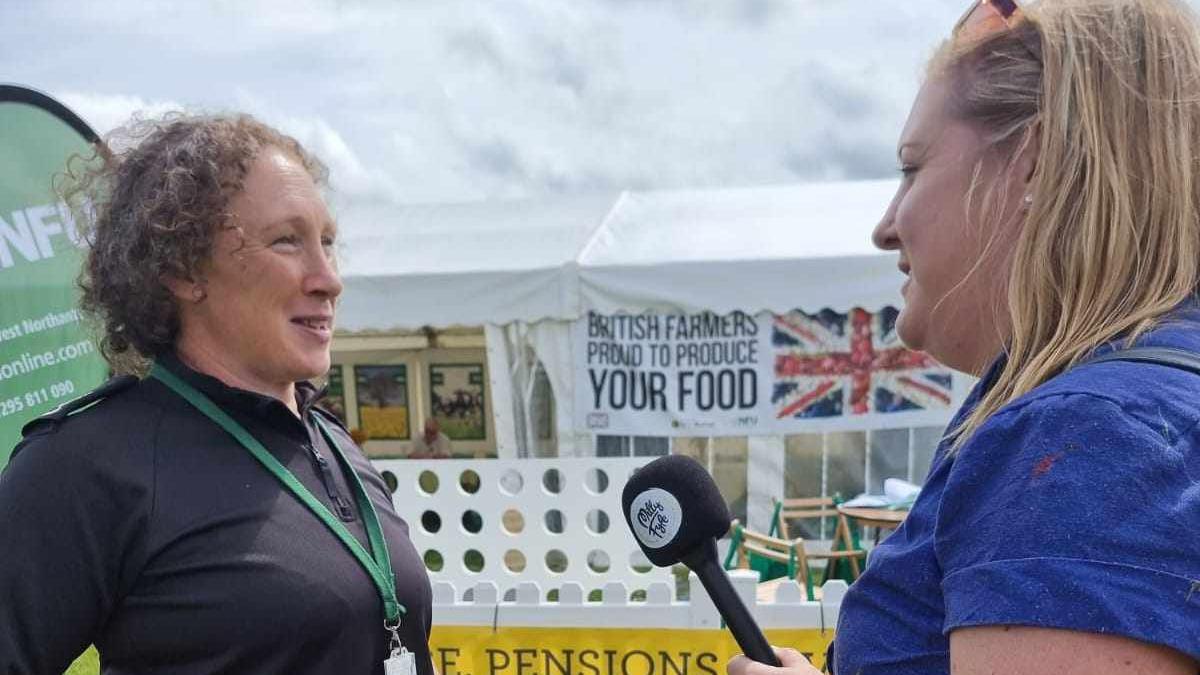 Milly Fyfe has long blond hair and is wearing a blue jacket and holding a microphone. She is interviewing a woman with dark curly hair wearing a green lanyard in front of a British Farmers marquee at a show