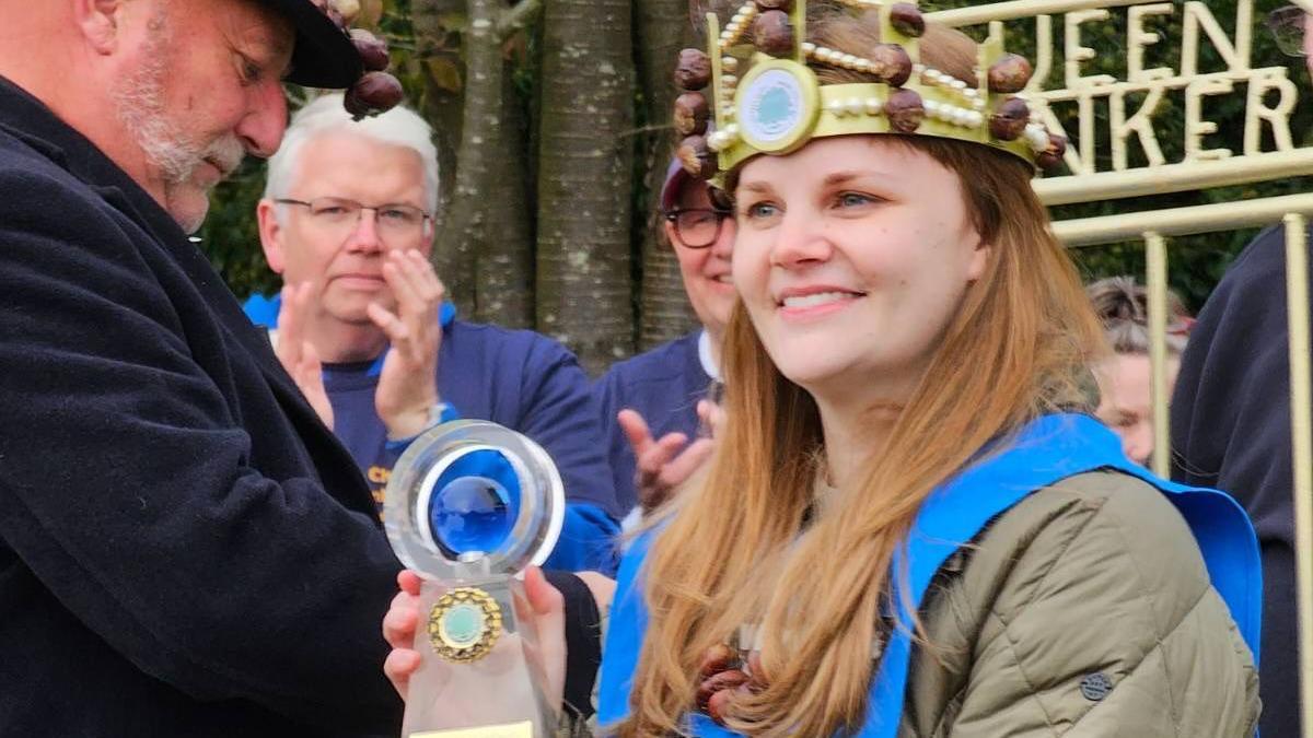 Kelci Banschbach has long light hair and is holding a glass trophy with a conker shape inside it. She also has a crown with conkers stuck to it. She is smiling as people applaud around her.