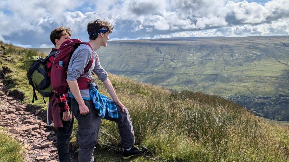 Arlo and another young man stand at the edge of a mountain taking in the scenery. Arlo wears a long sleeved grey sweatshirt, with charcoal grey cargo pants and hiking trainers. He has a blue checked shirt tied around his waist, a headband around his forehead - keeping his brown hair out of his eyes and is carrying a dark red hiking rucksak on his back. He is standing, resting his hand on his left leg surrounded by green mountains.