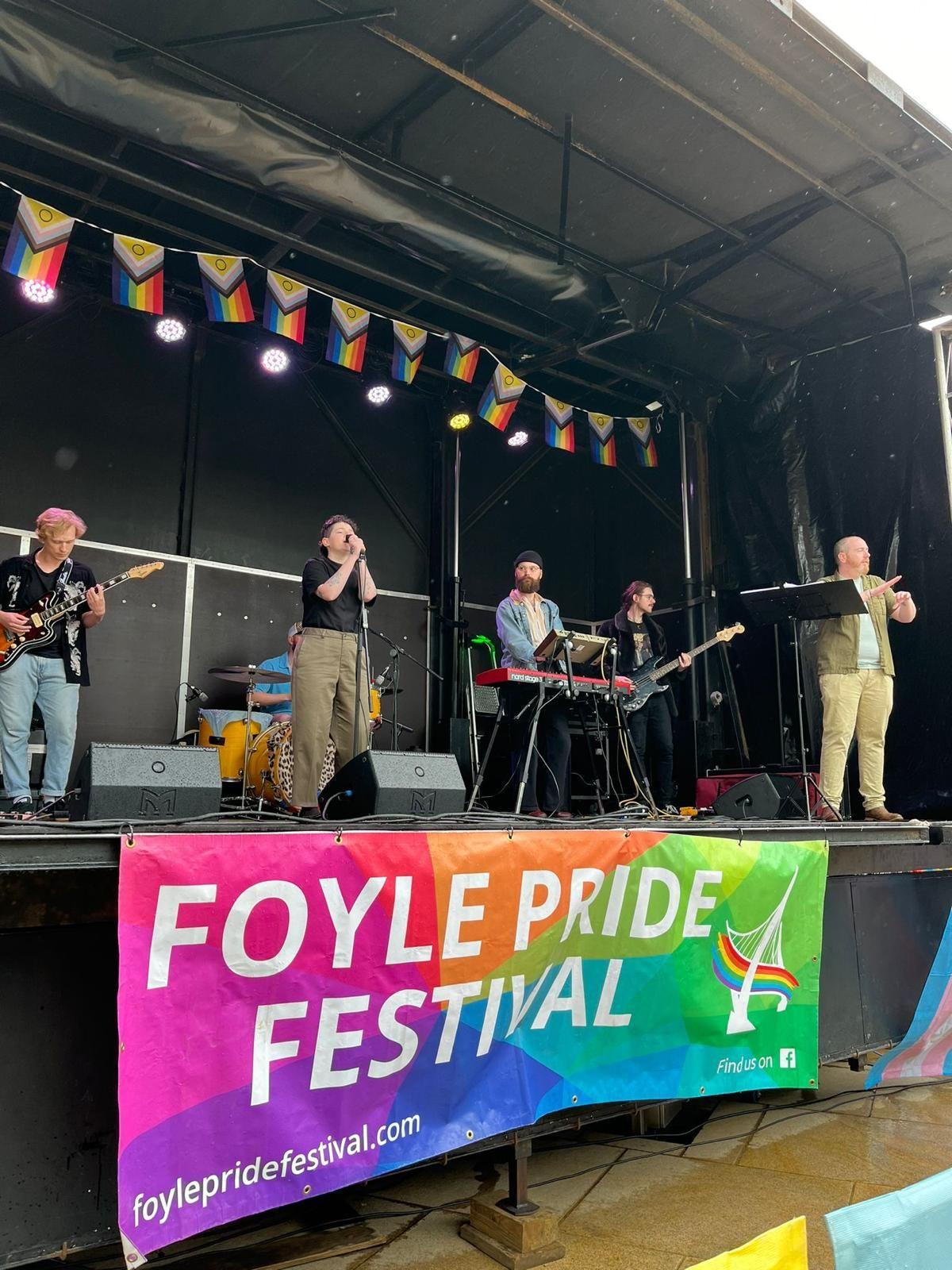 Five musicians on a raised stage with a large Foyle Pride banner