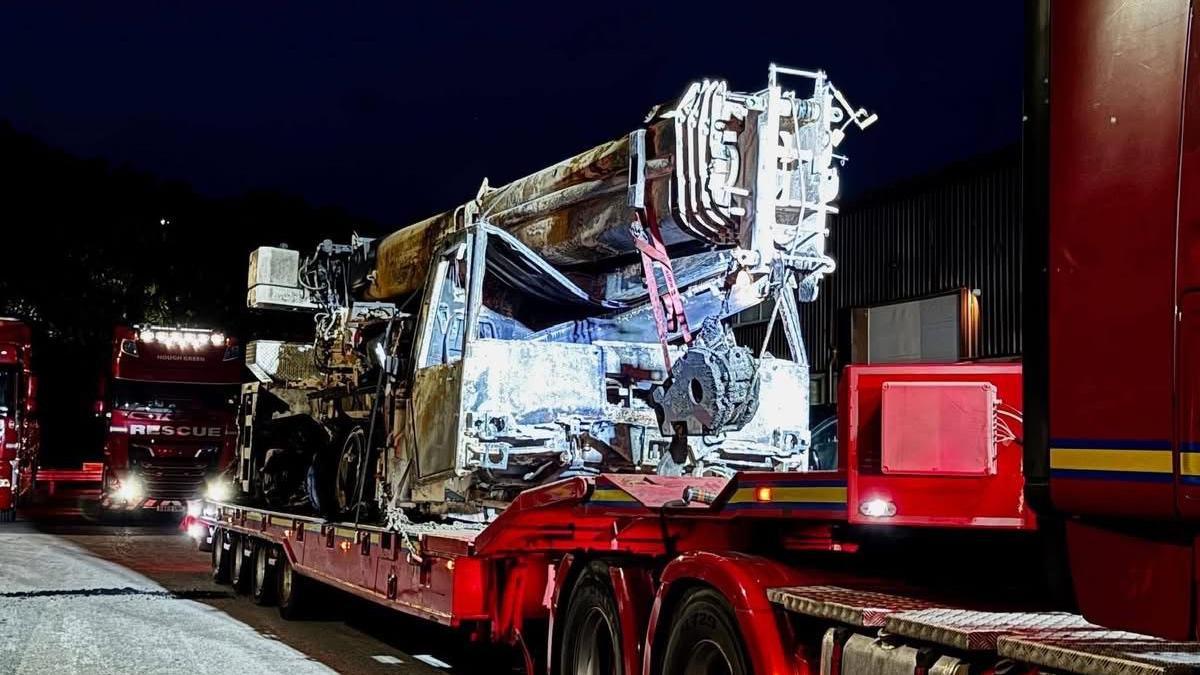 The damaged crane, lit up at night on the back of a red lorry. The crane is white and you can see fire-damage.