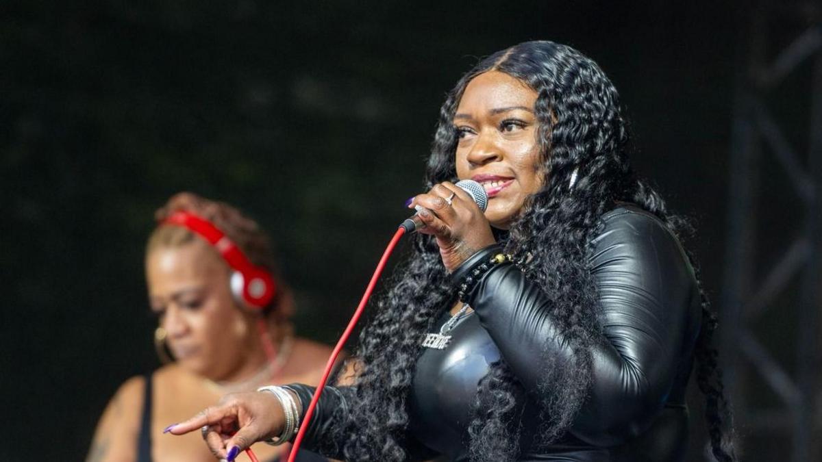 A woman with long crimped black hair sings into a microphone while pointing one finger toward the crowd. Another woman with red headphones can be seen behind her looking down.