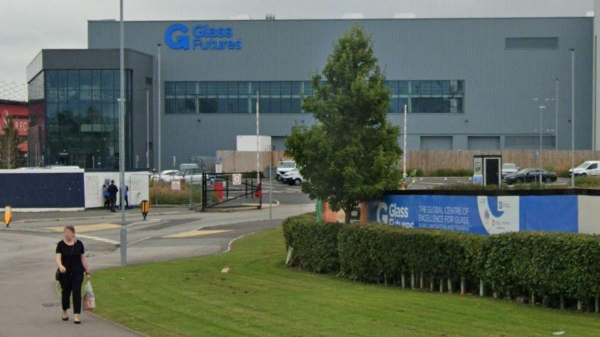 A large grey building with a glass frontage is set behind grass and a tree. There is blue signage which reads Glass Futures. A woman in in the foreground walking along with a shopping bag.