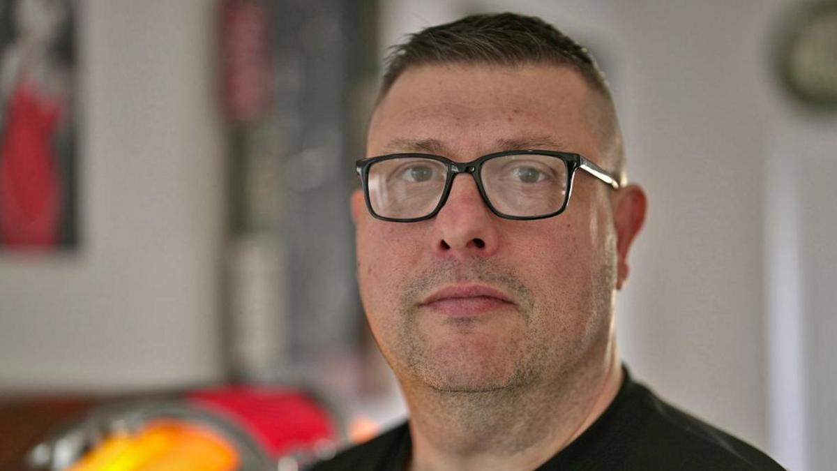 Brian Buckle looks straight towards the camera - he is wearing square glasses and a black jumper. He has short dark hair and dark stubble, and is pictured in a kitchen.