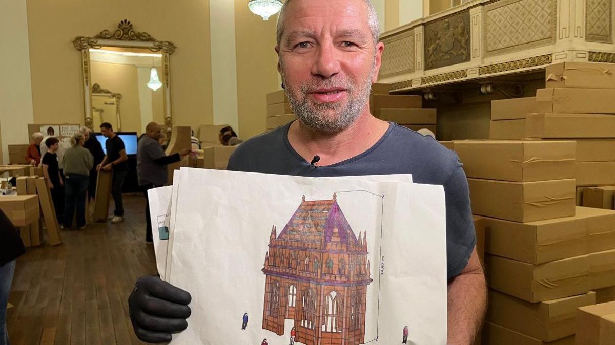 Jean-Marie Bergey stands with a plan of the structure being created, which has been inspired by the ruins of the bombed cathedral. The design on a plain sheet of paper, shows a building with a pointed roof, turrets and arched windows.