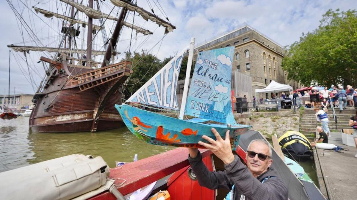 A man with white hair and sunglasses holding up a blue handmade model sailboat in front of a large wooden galleon ship. The model sailboat has a mermaid painted on the bottom and writing on its sails which reads 'The Lilarlic 2.0 will never sink - never'.