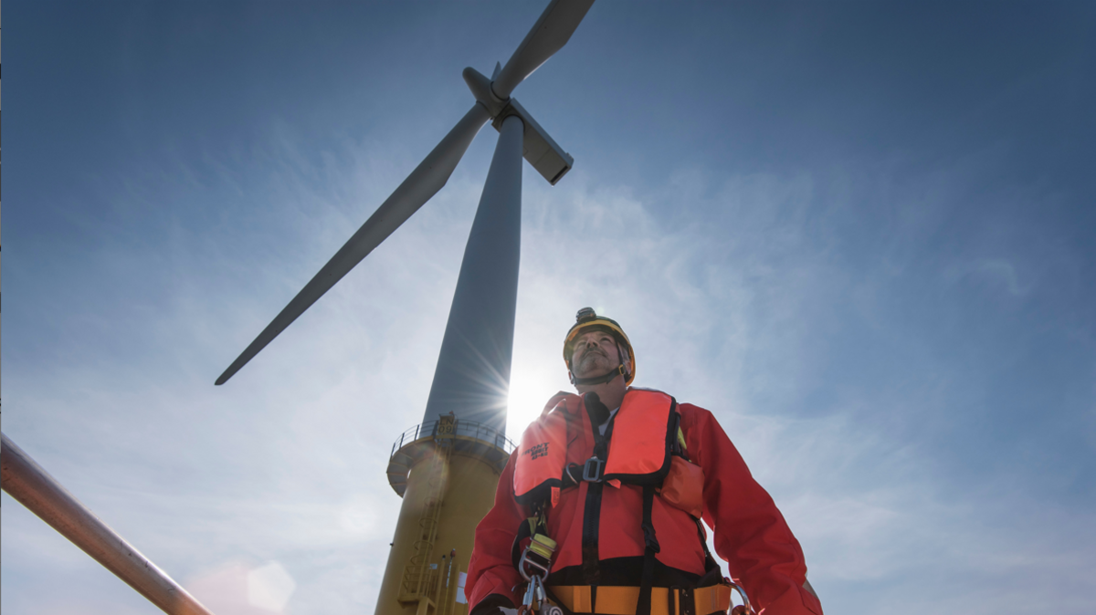 Male worker in florescent orange workwear and yellow helmet stands in front of an offshore wind turbine