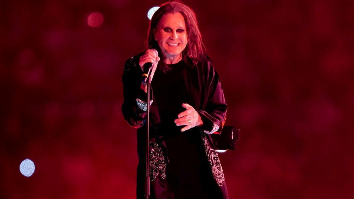 Ozzy Osbourne smiling with a microphone on a stand in his hand. He is wearing a long dark jacket and the background is red with some lights. 