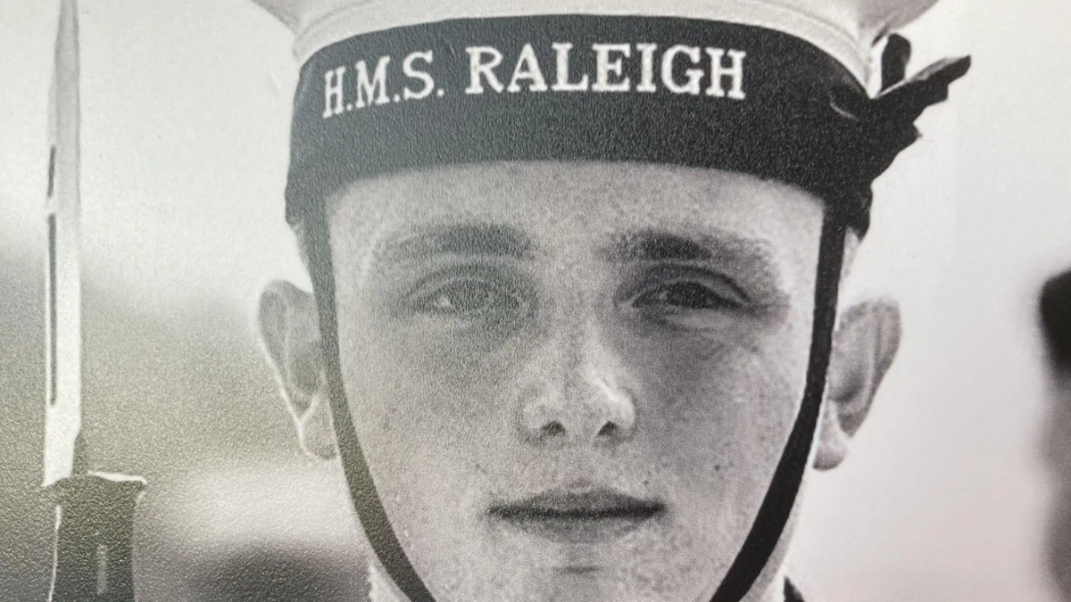 A black and white image of Joe Baker, wearing a H.M.S Raleigh hat and holding a gun with a bayonet 