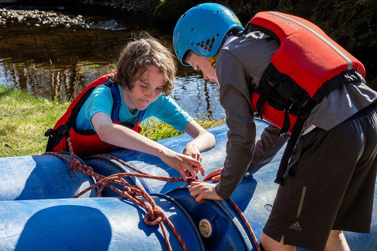 Two young boys in life jackets, one with a blue safety helmet on, lean over a raft they are constructing out of large blue plastic barrels, tying an orange rope to pull them together. A stream runs in the background