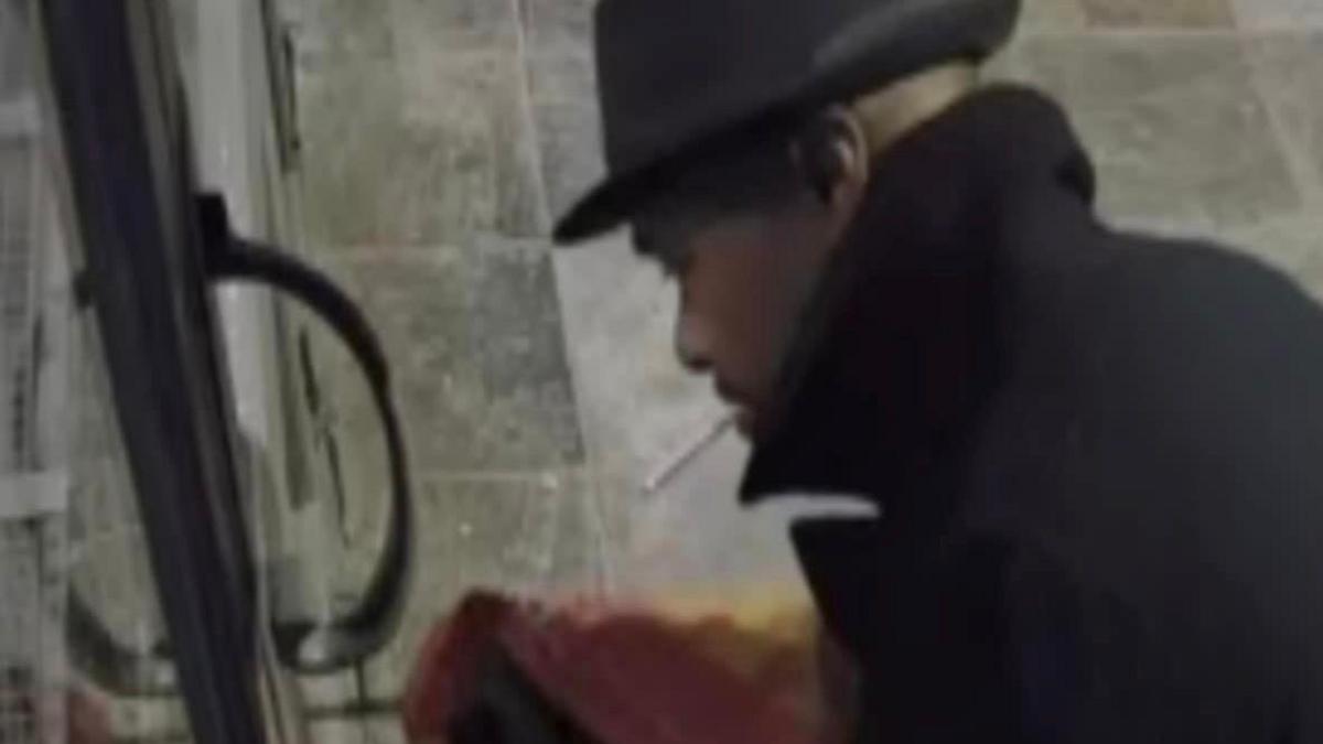 A CCTV image of a man wearing a black coat with the collar pulled up around his face. He has a cigarette hanging out of his mouth.