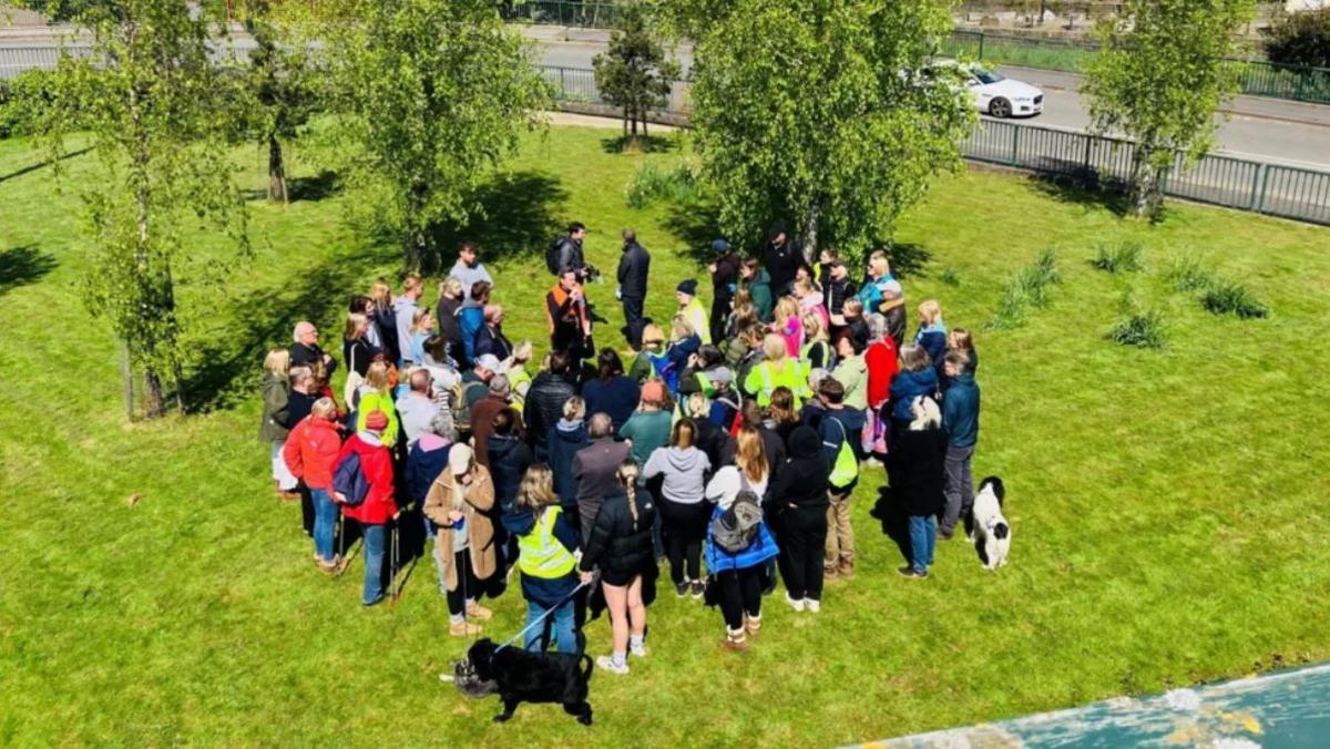 A large group of people gathered on a grassy patch beside the road. Some are wearing hi-vis vests and they are all listening to a group leader