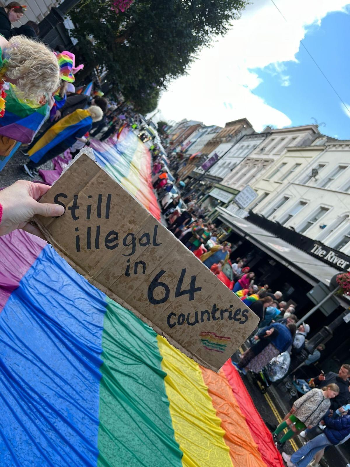 a hand holding a "still illegal in 64 countries" sign in front of a rainbow flag