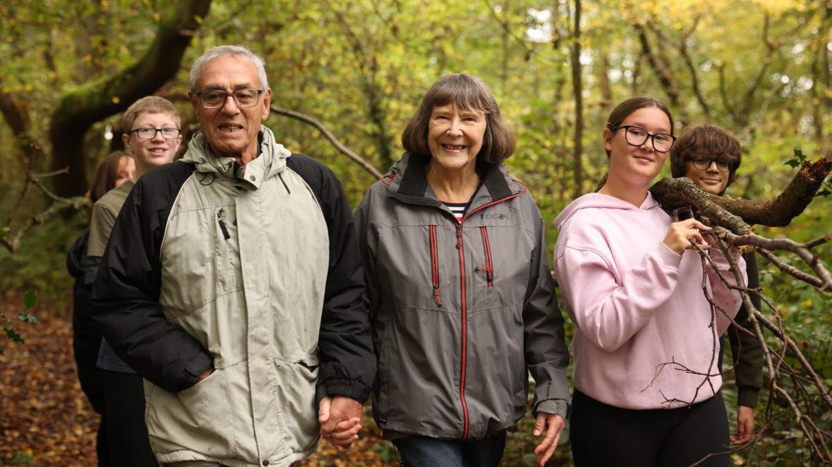 An older couple wearing cagoules walk hand in hand in the woods surrounded by teenagers. 