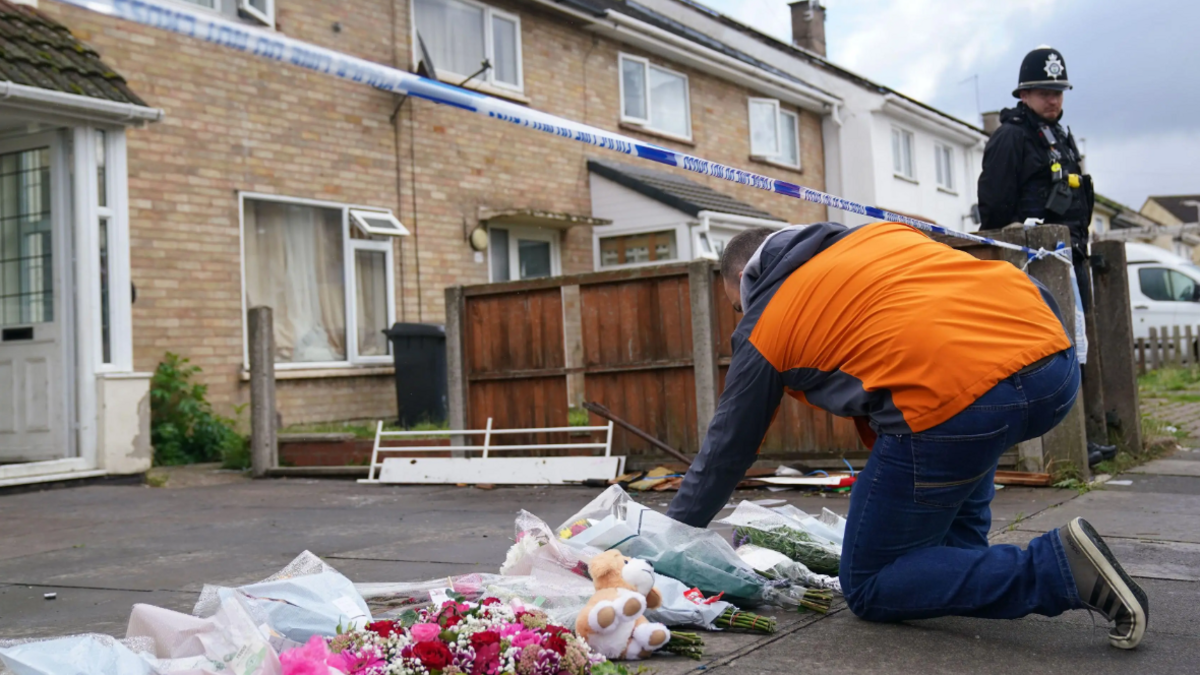 A man in an orange coat and jeans placing flowers on paving slabs outside a light brick house with police tape outsider and a police officer in full black uniform stood near by