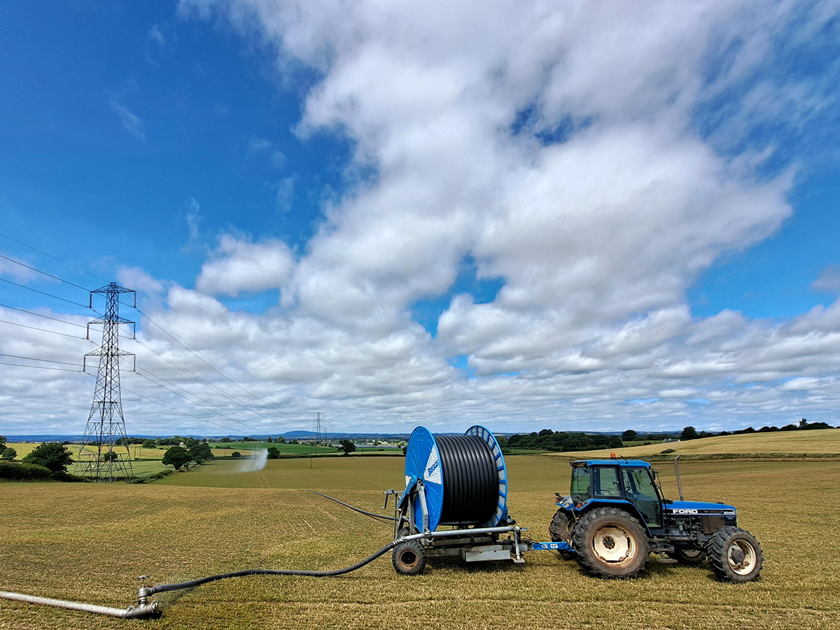 A wide view of a farm field near Wolverhampton. A blue tractor on the right is towing a roll of industrial hose. The black hose runs down the field to spray equipment irrigating plants. An electricity pylon is on the left and there are other green fields in the distance