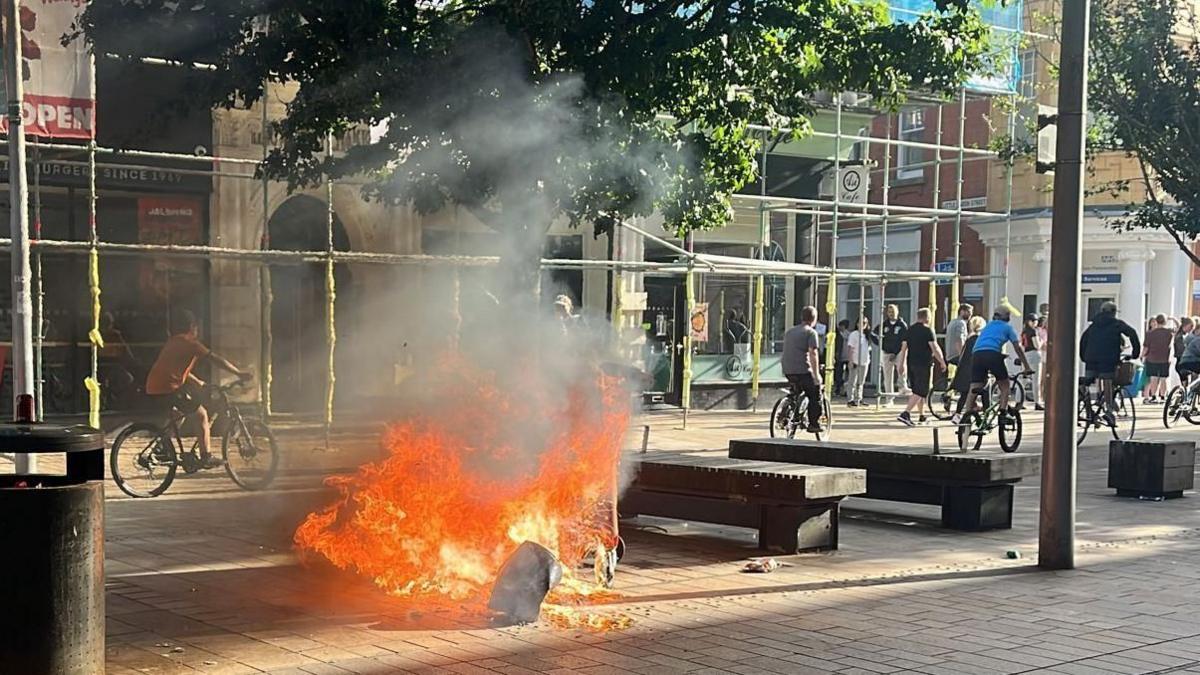 A fire burning from an upturned bin with people riding on their bikes in the background