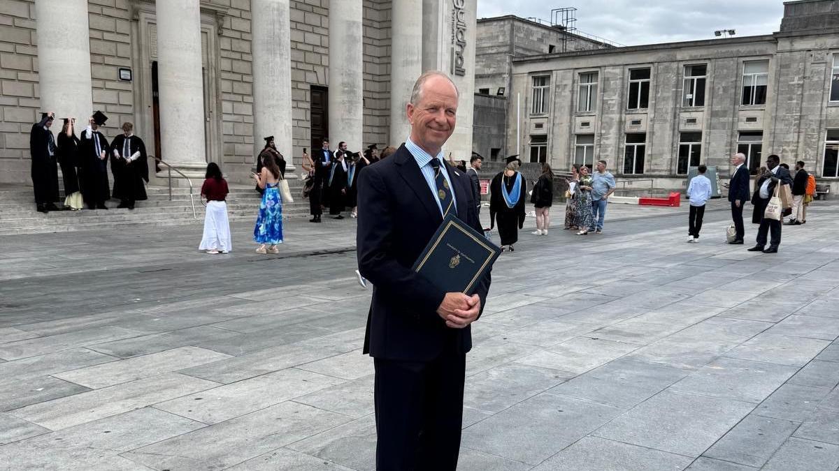 A man is stood in a blue shirt with a black blazer on top. He has a black tie with gold detailing on it. He is holding a certificate. He is smiling looking away from the camera. Behind him is the Guildhall.
