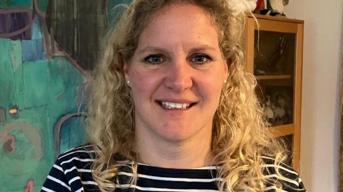 Camilla Notley has curly blonde hair and is smiling, looking at the camera. She is standing in front of one of Malcolm Race's abstract paintings. It has teal and turquoise shapes with grey lines.