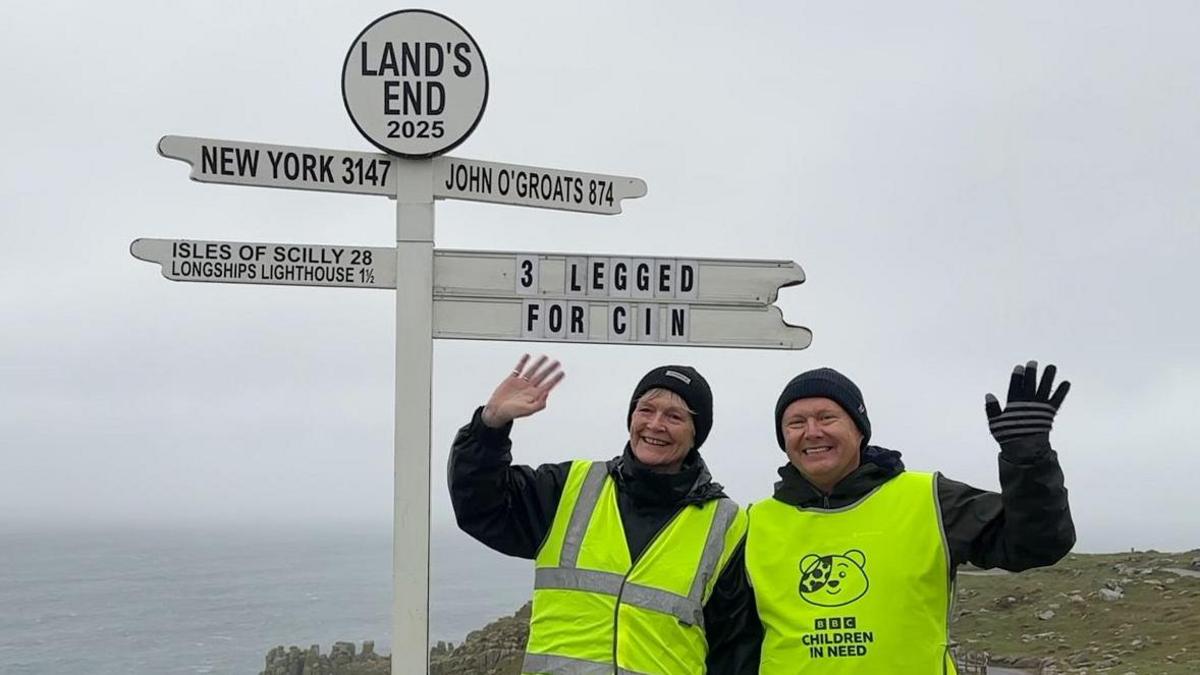 The photo shows radio presenter James Churchfield with a guest at the famous Land's End sign. They are both wearing fluorescent yellow Children in Need branded clothing and are waving at the camera. 