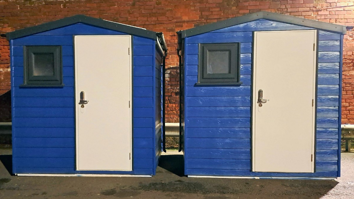 Two garden shed-type structures, painted blue and each with a white door and a small window, stand in front of a brick wall.