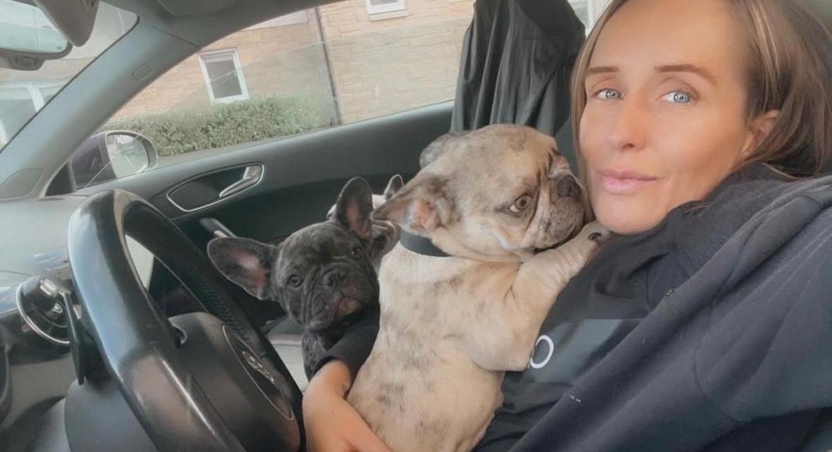 A woman with blonde hair sits in a car with two French bulldogs