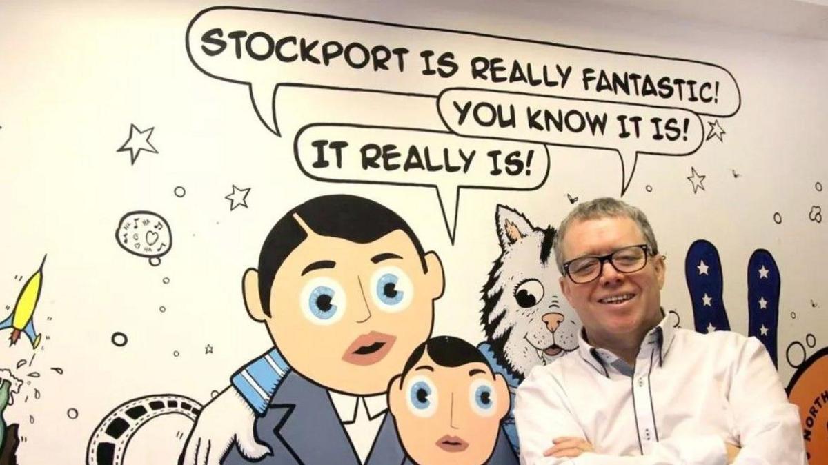 John Barratt, who is wearing a white shirt, is standing in front of a white wall which has been painted with pictures of people and animals. There is a speech bubble at the top which reads: "Stockport is really fantastic. You know it! It really is." John has his arms crossed and is smiling at the camera. 
