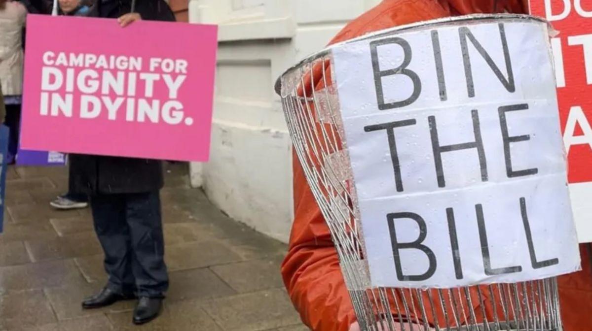 A pink sign that says Campaign for Dignity in Dying in white writing is held up by someone in dark clothing and shoes. Alongside that is a metal bin which has a white sign that says Bin the Bill bin attached to it, which is covered in raindrops and is being held by someone wearing a burnt orange raincoat. They are both standing in front of the white wall of the parliament building and the ground is wet will rainfall.