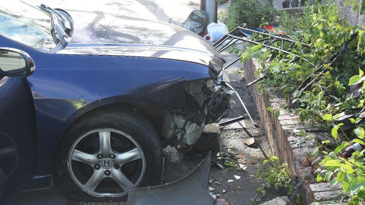 A dark blue Honda Accord. The front bumper is smashed up after being driven at a man. The car is on the pavement in front of a wall. A metal fence which was attached is lying on its side. 