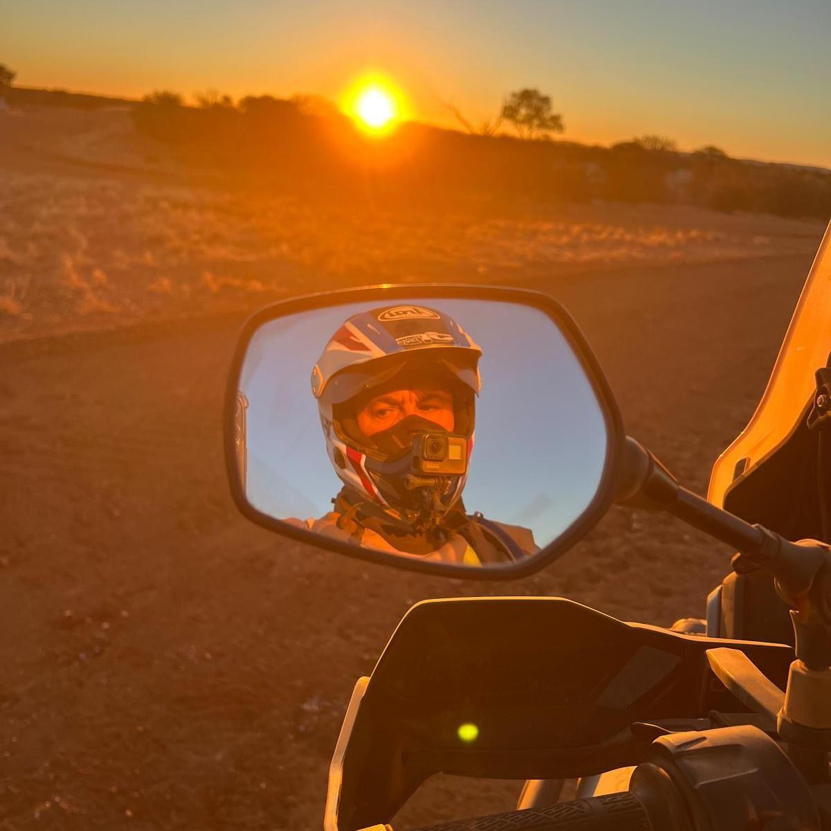 A picture taken from a motorbike, looking at the bike's rear-view mirror which shows the driver's face and helmet as he looks into the mirror. Beyond the road, the sun is setting over the horizon.