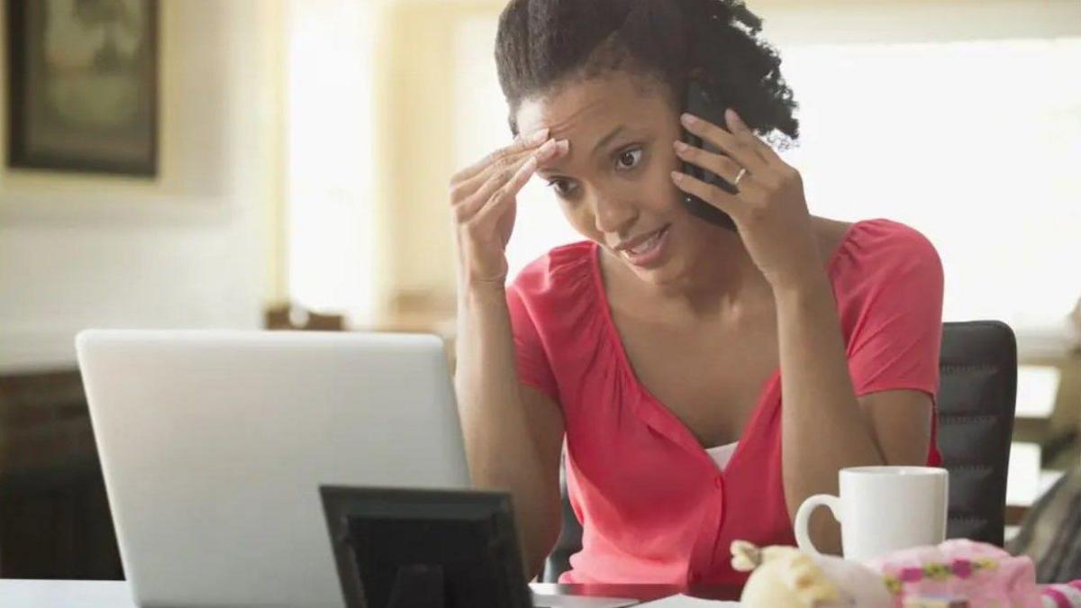 A woman in a red top speaking into a mobile phone in her left hand. She is sitting at a table in front of a laptop.