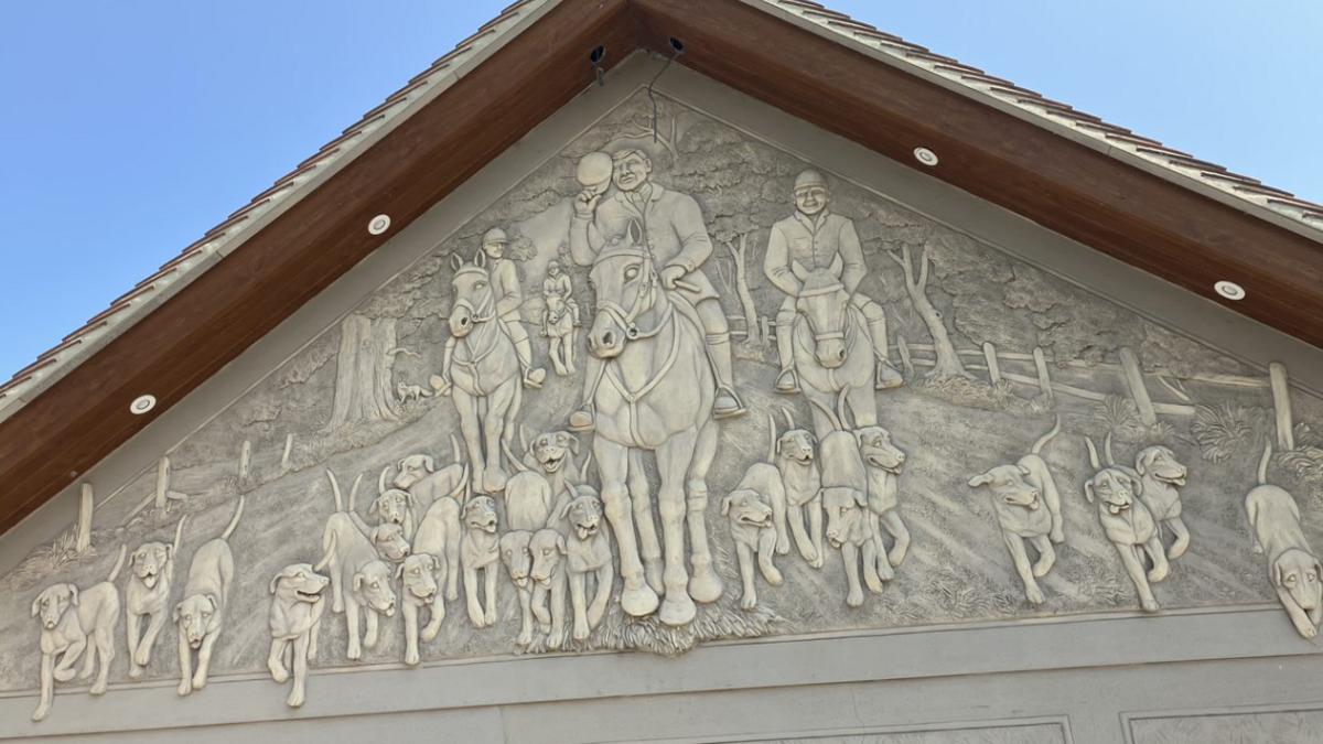 A scene of a pack of about 20 hunting dogs and four men on horseback, all walking towards us, created by pargeting on the side of a building, directly below the apex of its roof.