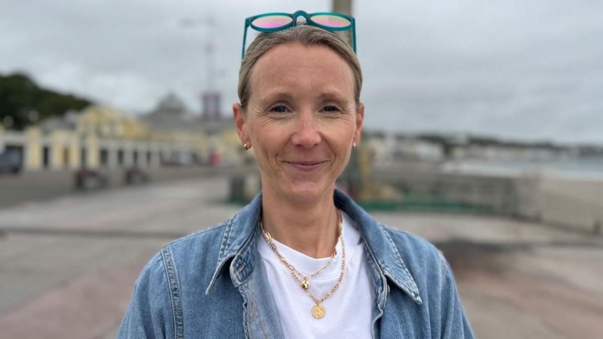 Lorna Gleave has fair hair tied back, with sunglasses resting on her head. She's wearing a white t-shirt and a denim jacket.